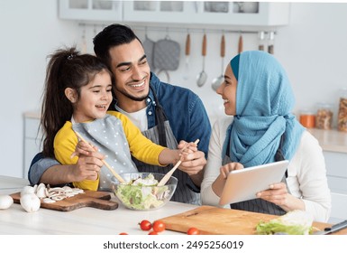 Happy Islamic Family Of Three Checking Food Recipe On Digital Tablet While Cooking In Kitchen Together, Cheerful Muslim Mom, Dad And Little Daughter Enjoying Preparing Lunch At Home, Closeup - Powered by Shutterstock