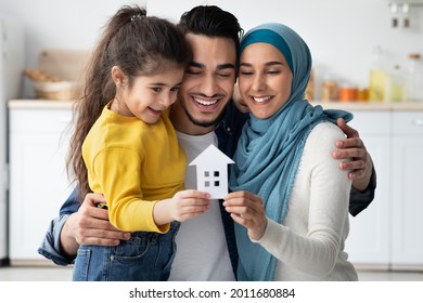 Happy Islamic Family Of Three With Little Daughter Holding Cutout Paper House Figure In Hands, Joyful Smiling Arab Parents And Cute Female Child Celebrating Buying New Property, Closeup Shot