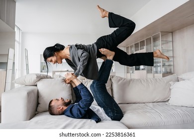 Happy Italian beardy man in casual laying on couch lifting up girlfriend by legs celebrating new apartment. Excited young hispanic couple having fun at hotel room on vacations. Newlyweds at honeymoon.