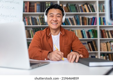 Happy Japanese Middle Aged Male Teacher Having Distance Class Online Video Calling On Laptop, Posing Smiling To Camera Wearing Headphones And Taking Notes Sitting At Workpace Indoor