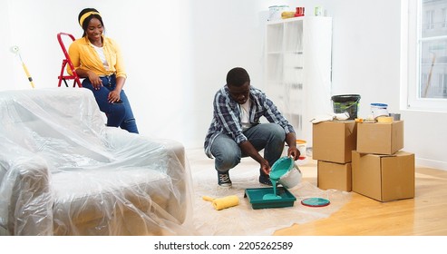 Happy Joyful African American Young Couple Doing Renovation In Room, Husband Pouring Green Paint Preparing For Painting Walls In New Color, Improving And Redecorating House, Renewing Home