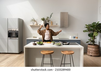 Happy joyful African dancer girl listening to music  dancing while preparing dinner in home kitchen  having fun at cooking island table and cut vegetables  healthy organic food ingredients