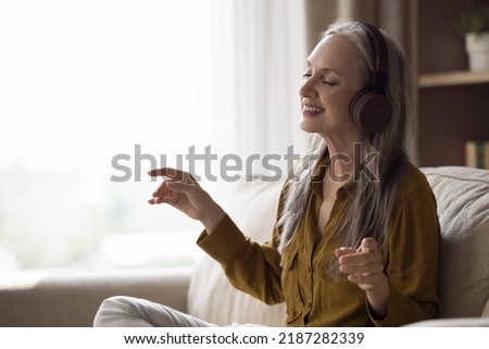 Happy joyful beautiful elderly lady in modern headphones having fun at home, listening to hifi music, favorite songs, singing along with closed eyes, sitting in comfortable couch