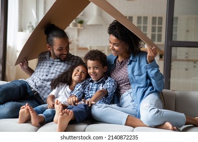 Happy Joyful Black Millennial Couple And Little Sibling Kids Having Fun, Playing Together On Couch, Holding Carton Roof, Symbol Of House, Safety Insurance. Homeowners Family Enjoying New Home