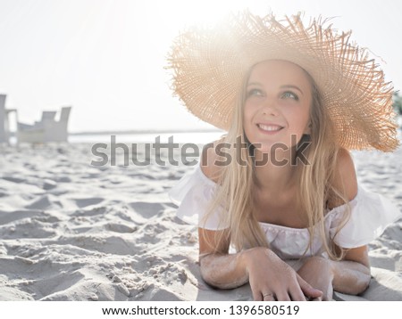 Happy and joyful blond young woman in white flounced top and a straw hat lays on a beach white sand enjoying the summer and morning sun. Summer vacations or holidays concept. 