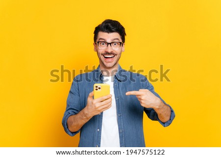 Happy joyful caucasian stylish guy with glasses, holding smartphone in hand and pointing at it finger, looking happily at camera with smiling, standing on isolated orange background