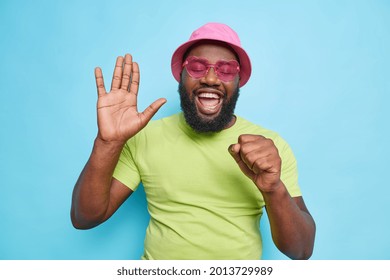 Happy joyful dark skinned man raises palm sings song keeps hand near mouth as if microphone has upbeat mood wears casual green t shirt pink panama trendy sunglasses isolated over blue background - Shutterstock ID 2013729989