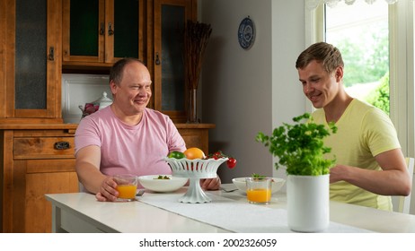 A Happy And Joyful Gay Couple Eating Pasta Together Drinking Orange Juice Enjoying The Afternoon In A White Kitchen With White Bowls And A White Fruit Bowl With A Flower Plant.