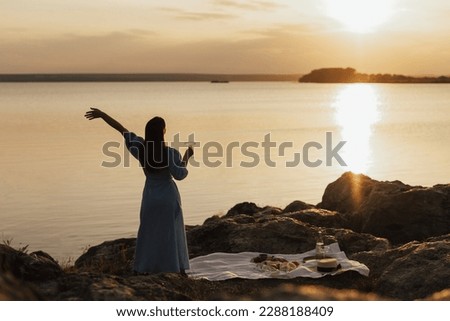 Happy joyful girl in long dress with hand up enjoy view and beautiful sunset at summer picnic. View from back. Copy space.
