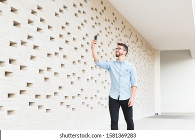 Happy joyful guy with smartphone taking picture or selfie in contemporary interior. Handsome young man in casual denim shirt standing indoors at modern brick wall. Taking picture concept - Powered by Shutterstock