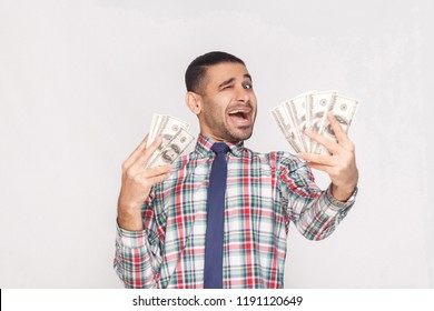 Happy joyful handsome young adult businessman in colorful checkered shirt with blue tie standing, holding fan of dollars and winking with open mouth. Indoor, studio shot, isolated on grey background