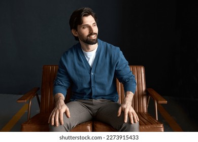 Happy joyful married guy with breaded chin having rest sitting on brown small couch, looking aside with toothy smile, putting hands on knees, wearing blue classical cardigan and stylish pants