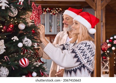 Happy Joyful Mature Older Couple Wearing Santa Hats Decorating Christmas Tree At Home Together Celebrating Festive Merry Xmas Holiday Party Eve, Feeling Joy In Cozy Wooden Winter House.