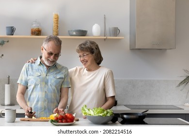 Happy Joyful Older Husband And Wife Sharing Cooking Chores, Making Organic Fresh Salad Together, Cutting Vegetables, Talking, Laughing. Old Couple Having Fun In Kitchen, Preparing Dinner