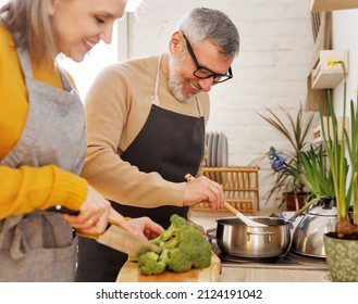 Happy joyful senior couple in aprons prepare vegetarian dinner in cozy kitchen, husband stirring food with wooden spoon in pan while wife cutting broccoli for cream soup. Cooking on retirement - Powered by Shutterstock