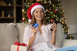 Happy Joyful Shopper Girl In Xmas Santa Hat Buying Christmas Gifts Online, Using Smartphone, Credit Card, Paying On Internet, Looking At Camera, Smiling, New Year Home Shopping. Head Shot Portrait