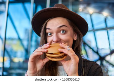 Happy Joyful Stylish Hipster Hungry Woman With Surprised Wide Eyes Eats Burger In Fast Food Restaurant