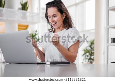 happy joyful successful remote working woman cheerful with fists infront of laptop or notebook in casual outfit sitting on work desk in modern loft living room home office