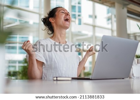happy joyful successful remote working woman cheerful with fists infront of laptop or notebook in casual outfit sitting on work desk in modern loft living room home office