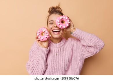 Happy joyful woman covers eye with delicious glazed doughnut laughs happily dressed in casual knitted sweater poses indoor against beige background has sweet tooth breaks diet. Junk food concept - Shutterstock ID 2099586076