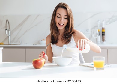 Happy joyful woman pouring milk into a bowl while sitting and having breakfast at the kitchen table - Powered by Shutterstock