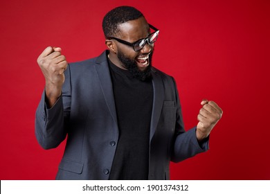 Happy Joyful Young African American Business Man 20s Wearing Classic Jacket Suit Eyeglasses Standing Doing Winner Gesture Clenching Fists Isolated On Bright Red Color Wall Background Studio Portrait