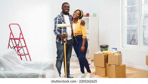 Happy joyful young African American married couple embracing standing in new apartment during home reconstruction with roller brush for painting wall looking at camera and smiling, renovation - Powered by Shutterstock