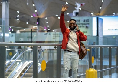 Happy Joyful Young African Man Traveler Waving Hand And Talking By Phone, Saying Hi Or Goodbye While Standing With Suitcase In Airport, Cheerful Black Guy Waiting Friends In Terminal. Travel Concept