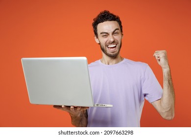 Happy Joyful Young Bearded Man 20s Wearing Casual Basic Violet T-shirt Standing Working On Laptop Pc Computer Doing Winner Gesture Isolated On Orange Background Studio Portrait. Tattoo Translate Fun