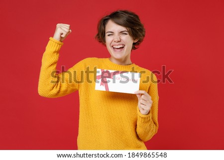 Happy joyful young brunette woman 20s wearing basic yellow sweater standing hold in hands gift certificate doing winner gesture looking camera isolated on bright red colour background studio portrait