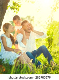 Happy Joyful Young Family Father, Mother And Little Son Having Fun Outdoors, Playing Together In Summer Park. Mom, Dad And Kid Laughing And Hugging, Enjoying Nature Outside. Sunny Day, Good Mood