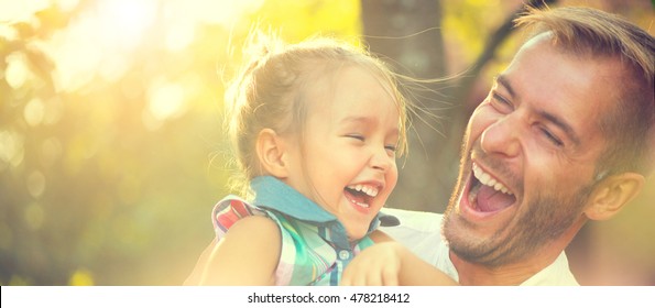 Happy Joyful Young Father With His Little Daughter. Father And Little Kid Having Fun Outdoors In Orchard Garden, Playing Together In Summer Park. Dad With His Child Laughing And Enjoying Nature