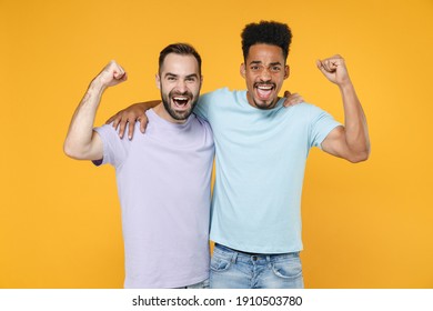 Happy joyful young friends european african american men 20s in casual violet blue t-shirts hugging doing winner gesture looking camera isolated on bright yellow colour background studio portrait