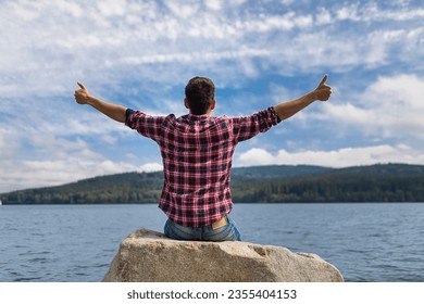 Happy Joyful Young Man Feeling Alive Joyful Positive Thumbs Up Sitting Next to a Beautiful Lake Enjoying Peaceful Nature
