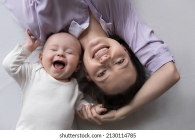 Happy joyful young mother and adorable few month baby resting together on big bed mattress with white linen, laughing, smiling, looking at camera. New mom with little kid home portrait. Top view
