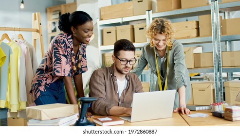 Happy joyful young multi-ethnic co-workers speaking while browsing online searching internet on laptop. People at work in clothing manufacture store, fashion style designer, shop warehouse concept