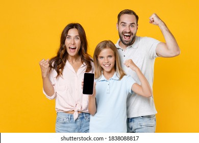 Happy Joyful Young Parents Mom Dad With Child Kid Daughter Teen Girl In Basic T-shirts Hold Mobile Phone With Blank Empty Screen Doing Winner Gesture Isolated On Yellow Background. Family Day Concept