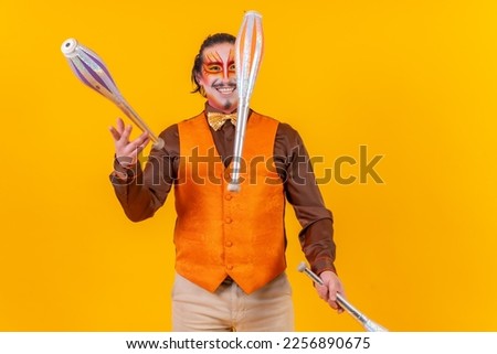 Happy juggler man in makeup vest juggling maces on a yellow background