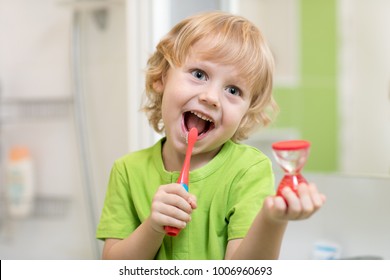 Happy Kid Boy Brushing Teeth Near Mirror In Bathroom. He Is Monitoring Lasting Of Cleaning Action With Hourglass.