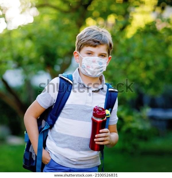 Happy kid boy, medical mask, water bottle and\
backpack or satchel. Schoolkid on way to school. Healthy child\
outdoors. Back to school after quarantine time from corona pandemic\
disease lockdown