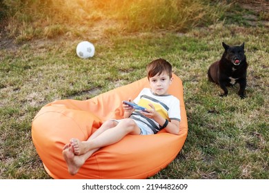 Happy Kid Boy Playing Game On Smartphone In The Park Outdoor, Child Using Smartphone On Soft Orange Chair At Home Garden After Active Games With Ball And Dog, Backyard, Sunlight, Cell Phone Addiction