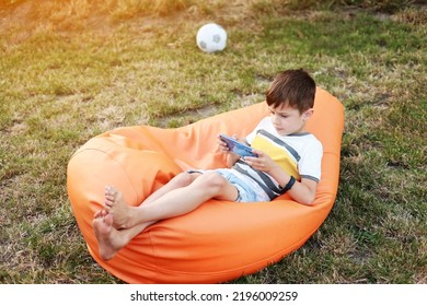 Happy Kid Boy Playing Game On Smartphone In The Park Outdoor, Child Using Smartphone On Soft Orange Chair At Home Garden After Active Games In Football, Backyard, Sunlight, Cell Phone Addiction