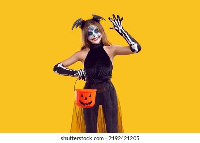 Happy Kid Celebrating Halloween And Going Trick Or Treating. Portrait Of Child In Spooky Costume Isolated On Yellow Background. Little Skeleton Girl Holding Her Candy Basket, Waving Hello And