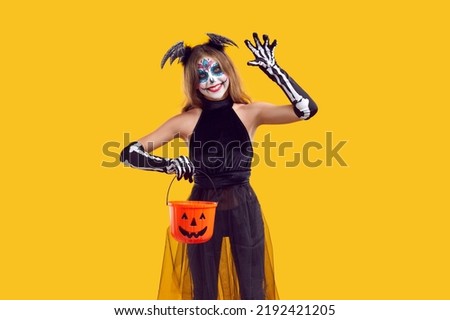 Happy kid celebrating Halloween and going trick or treating. Portrait of child in spooky costume isolated on yellow background. Little skeleton girl holding her candy basket, waving hello and
