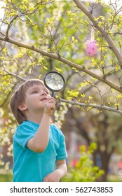 Happy Kid Enjoying Sunny Late Spring Easter Day And In Nature Hunting For Eggs With Magnifying Glass