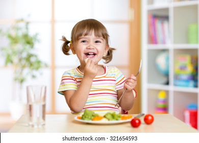 Happy Kid Girl Eating Healthy Food Vegetables At Home In Nursery