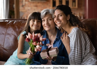 Happy Kid Girl, Mom, Grandma Celebrating 8 March, Birthday, Holding Bunch Of Flowers, Tulips, Gift Box, Sitting On Couch At Home, Hugging, Looking At Camera, Smiling. Three Family Generations Portrait