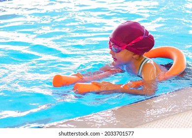 Happy  Kid Girl  At Swimming Pool Class Learning To Swim. The Girl Has Pink Hat, Swimming Glasses And A Noodle. Development Of Children's Sports, Strengthening Baby's Health.