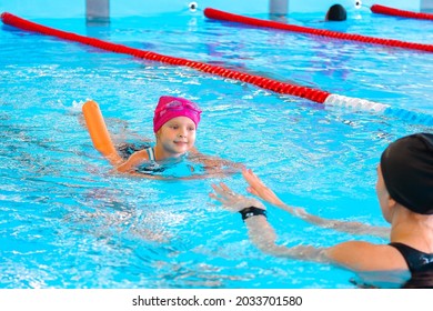 Happy  Kid Girl  At Swimming Pool Class Learning To Swim. The Girl Has Pink Hat, Swimming Glasses And A Noodle. Development Of Children's Sports, Strengthening Baby's Health.