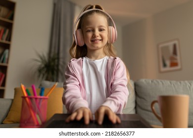 Happy Kid Girl Typing On Laptop Wearing Pink Headphones Smiling To Camera At Home. Schoolgirl Browsing Internet And Learning Online Having Video Call Indoor. Knowledge And Technology